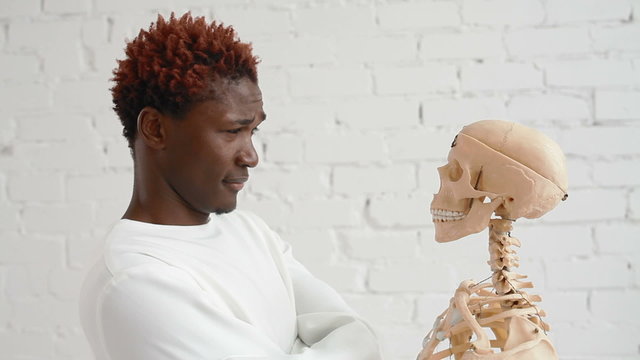 An insane black man wearing a straitjacket dance and looking at human skeleton stand
