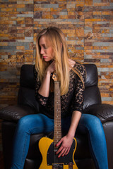 Fototapeta na wymiar Attractive young girl in blue jeans and black lace shirt sitting in chair and holding guitar on brick background 