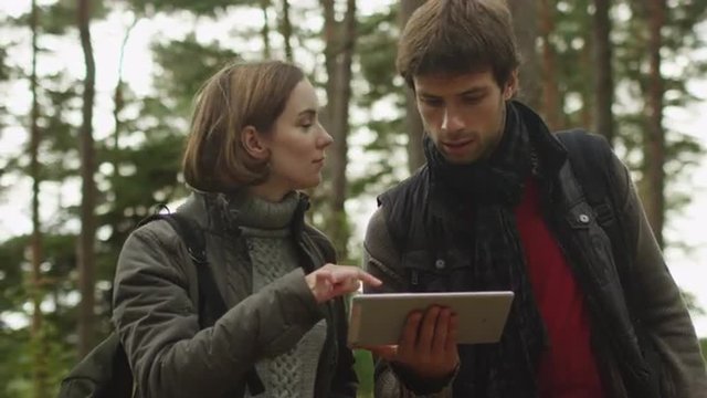 Long haired man and brunette woman in dark autumn clothes are standing in a forest and using tablet. Shot on RED Cinema Camera.