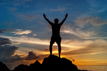 Fototapeta premium Man standing on the rock of a tropical island with his hands up and watching the sunset