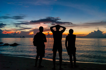 Three men standing on the beach of a tropical island  and  watching the sunset