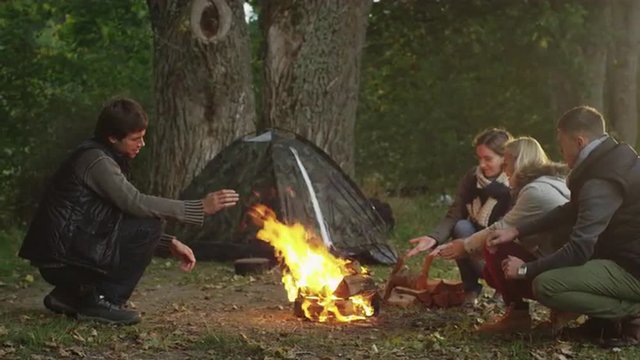 Group of people are sitting next to a campfire next to a tent in an autumn forest.
