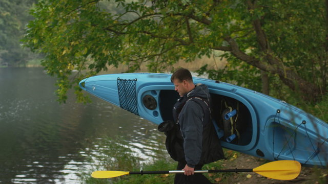 Man is carrying a blue kayak to the river shore and puts it on water