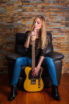      Attractive young girl in blue jeans and black lace shirt sitting in chair and holding guitar on brick background 