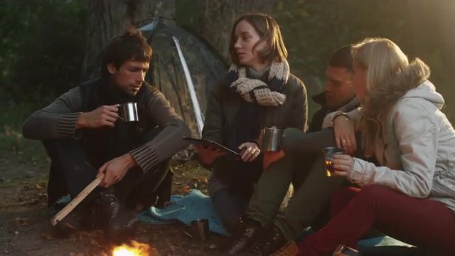 Group of people sit in a forest next to a campfire with warm drinks from thermos and use tablet. Shot on RED Cinema Camera.