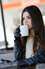 Woman with a unique fleshy lips wearing leather jacket and gloves holding a coffee cup, sitting in cafeteria