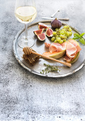 Wine appetizer set. Glass of white wine, honeycomb with drizzlier, figs, grapes, melon and prosciutto on silver tray over rustic grunge surface. 