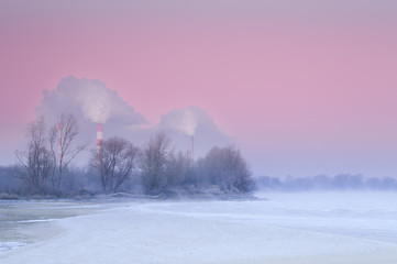 Smoking chimneys over a misty and freezing river during dusk