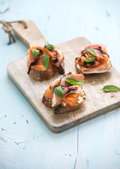 Bruschettas with Prosciutto, roasted melon, soft cheese and basil on wooden serving board over light blue background.