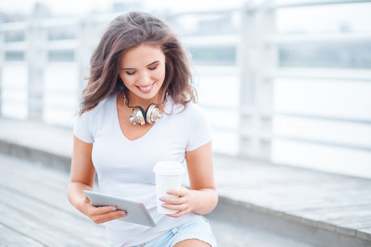 Music near water / Happy young woman with music headphones and a take away coffee cup, surfing internet on tablet pc, listening to the music and sitting on the bridge.
