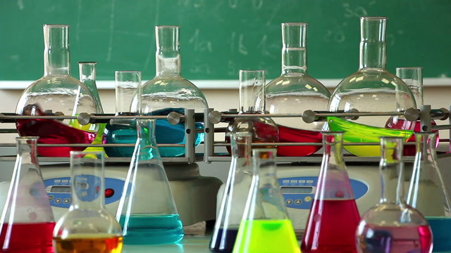 Laboratory shakers agitate eight round glass flasks filled with color solutions on the green chalkboard background. Other glasswares stand in front. Focus on the both shakers. No camera movement.
