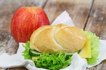 Lunch consisting of cheese roll with lettuce and red apple on rustic wooden surface
