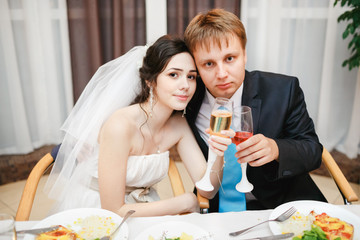 Newlyweds at the wedding table sitting together, posing with wine. Attractive beautiful bride, kind eyes, groom wearing suit and turquoise tie. Celebration, reception in restaurant is over.