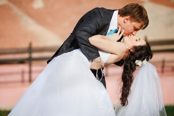 Groom gentle tilted bride, holding her in his arms and passionately kisses, wedding photo on a sunny day, background of sand-colored walls. Newly married couple dancing at park, street tango. 