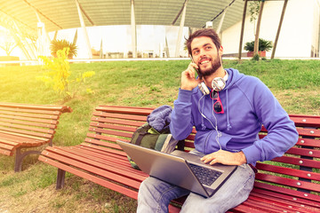 Young man using mobile phone and laptop pc - Guy with smartphone and laptop in urban park - Smiling...