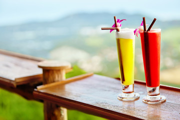 Drinks. Close Up Of Exotic Cocktails In Tall Glasses With Orchids, Drinking Straws On Table In Luxury Tropical Bar.  Beautiful Landscape ( View ) On Background. Thailand Vacations. Celebration.