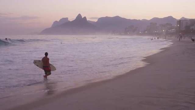 Slow motion pan of surfer at sunset along sandy beach with mountains and city in the background