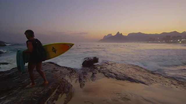 Slow motion, tracking shot of a surfer climbing rocks on Ipanema beach