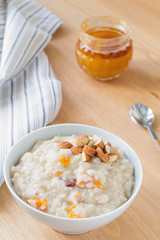 Oatmeal porridge with dried apricots and chopped almonds on bright wooden table. Jar of honey on background. Selective focus. Bright healthy breakfast image