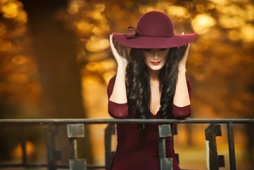 Attractive young woman with burgundy colored large hat in autumnal fashion shot. Beautiful mysterious lady covering the face with hat. Elegant brunette with gorgeous mouth, outdoors shot in fall.