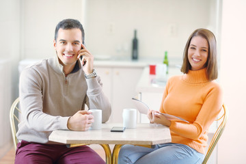 Happy young couple is having a coffee at home. Young man is talking at his mobile phone and young woman is reading a magazine
