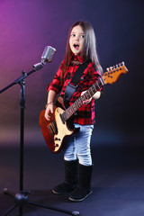 Obraz premium Beautiful little girl with microphone and guitar on dark purple background