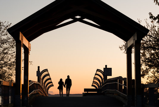 The silhouette of a couple holding hands walking into the sunset on a boardwalk.