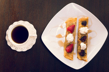 Two beautiful crepes pancakes lying on white plate, decorated with small amount of cream and berries, coffee cup next to it, elegant presentation