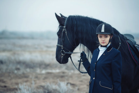 A Tall Girl With Long Blond Hair In A Jockey Outfit With A Beautiful Black Horse In An Empty Snow-covered Field In Winter