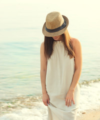 Happy smiling woman walking on a sea beach dressed in white dress and hat covering face, relaxing and enjoy fresh air.