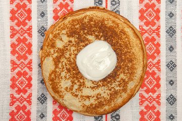 Pancakes and sour cream at tablecloth with patterns, top view