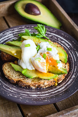Poached egg and avocado on toasted whole wheat bread with seeds on a plate in vintage wooden tray. Garnished with scallions and parsley.