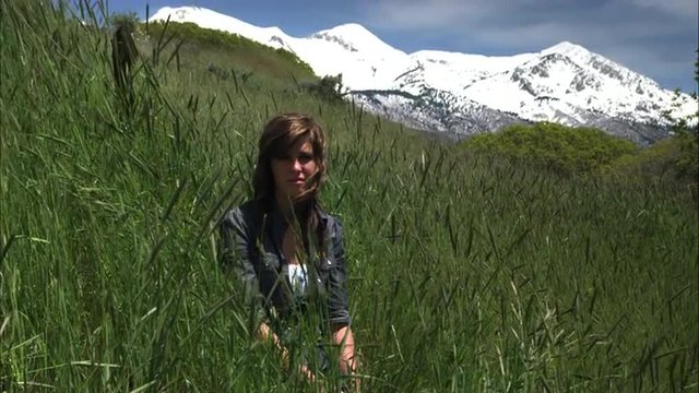 Slow motion shot of a young woman seated in tall grass with mountains behind her.