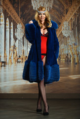 A young and attractive blonde woman wearing red lingerie, stockings and a fur coat standing in front of luxury background.