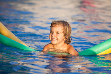 Fototapeta na wymiar happy child in the pool, learning to swim with swimming noodle; warm sunset light