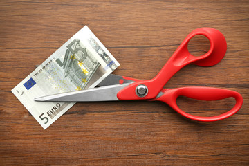 Red scissors cut money on wooden background. Financial concept