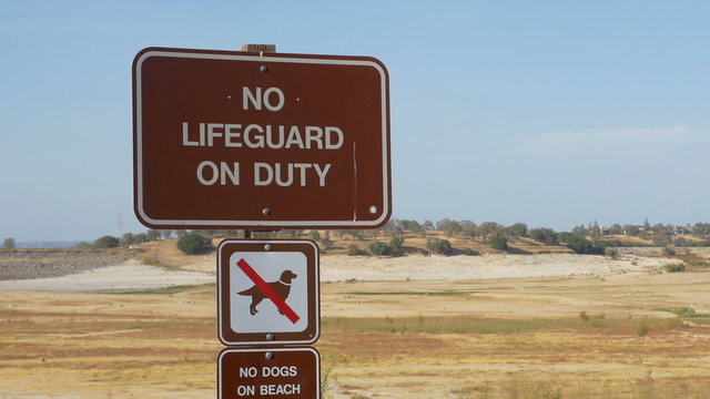 Zoom out of No Lifeguard On Duty sign, and pan right over a dry portion of Folsom Lake in October 2015.