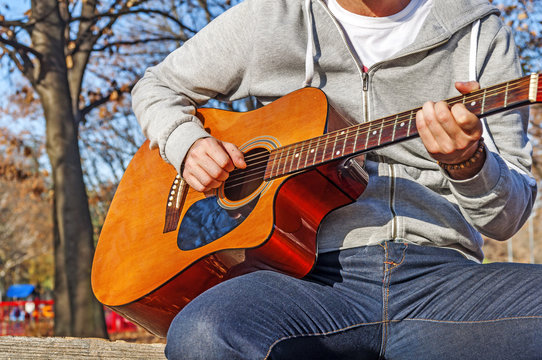 Young man sitting on wooden chair in park and playing guitar