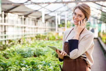 Smiling woman gardener using mobile phone and tablet in greenhouse