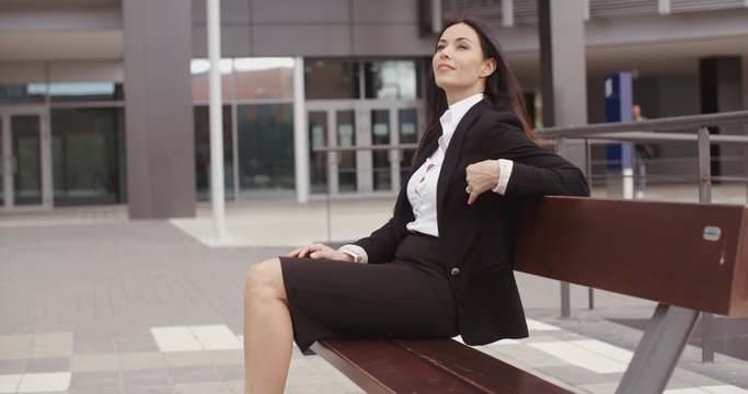 Confident young business woman sitting alone on bench outside office building with calm expression
