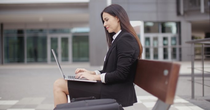 Cute business woman on bench in front of office building looking over while working on laptop computer