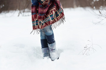 The figure of a young woman in bright ethnic scarf with purple, white and blue pattern, blue jeans and soft boots. She stands in the snow.