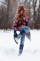 Young red-haired woman in the winter forest. She is dressed in an ethnic scarf with purple, white and blue pattern, blue jeans and soft boots. She has long red curly hair. She is dancing in the snow.