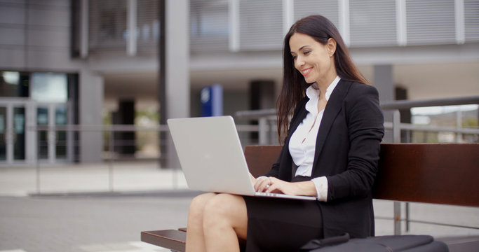 Cute business woman sitting alone on bench in front of office building working on laptop computer