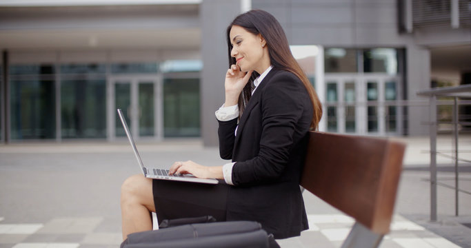 Cute business woman on bench in front of office building looking over while working on laptop computer