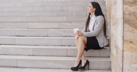 Grinning brunette professional woman sitting on outdoor stairs while drinking coffee