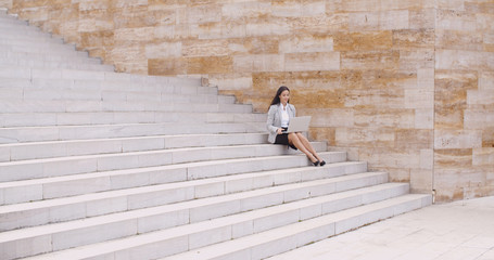 Cute business woman sitting alone on marble staircase outdoors with laptop from a distance