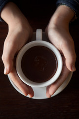 Female hands holding a cup of hot chocolate with foam over wooden table, top view 