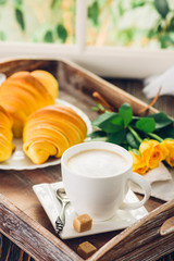 Coffee, croissant and flowers on tray next to the window