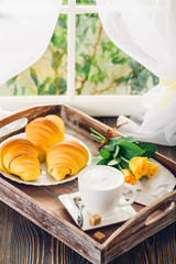 Fototapeta premium Coffee, croissant and flowers on tray next to the window